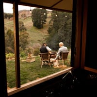Through the window you spot two people in chairs with their dog around a fire and the hills in the background 
