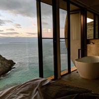 A image of a bath and floor-to-ceiling windows which look out over the ocean and cliffs