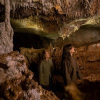 Two young women walk through a a cave, with a low rocky roof and rocky formations on either side of them