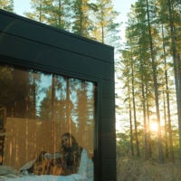Tall pine trees surround the cozy cabin where light shines inside, where a girl is reading her book. 
