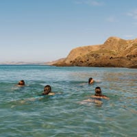 A group of girls swim in the water, a large cliff in the backdrop