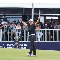 A woman holds up her golf club in celebration on a golf course with spectators behind her 